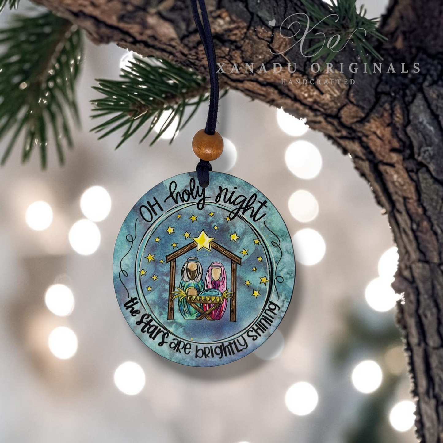 A round Christmas ornament hanging from a tree branch with a brown cord and wooden bead. The ornament features a watercolor-style illustration of the Holy Family under a starry night sky, with the text 'Oh holy night the stars are brightly shining.
