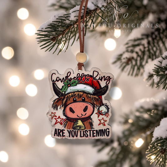 A wooden Christmas ornament shaped like a cow wearing a Santa hat hangs from a pine tree branch with snowy lights in the background. The ornament reads 'Cowboill Ring ARE YOU LISTENING' and has a warm, festive look.