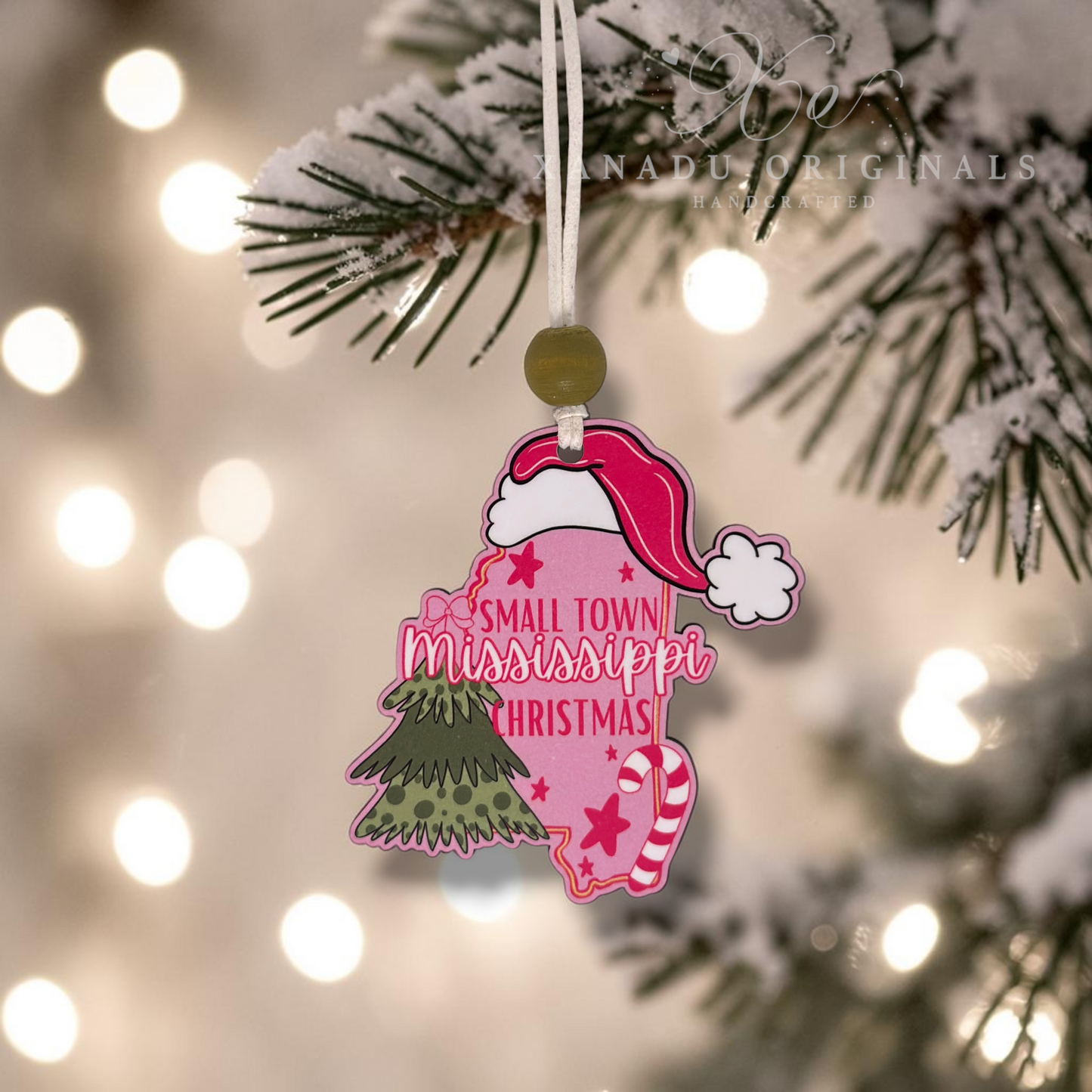 A small decorative Christmas ornament shaped like the state of Mississippi hangs from a snowy pine branch. The ornament is pink with a red Santa hat, white clouds, a green tree, and the words “Small Town Mississippi Christmas.
