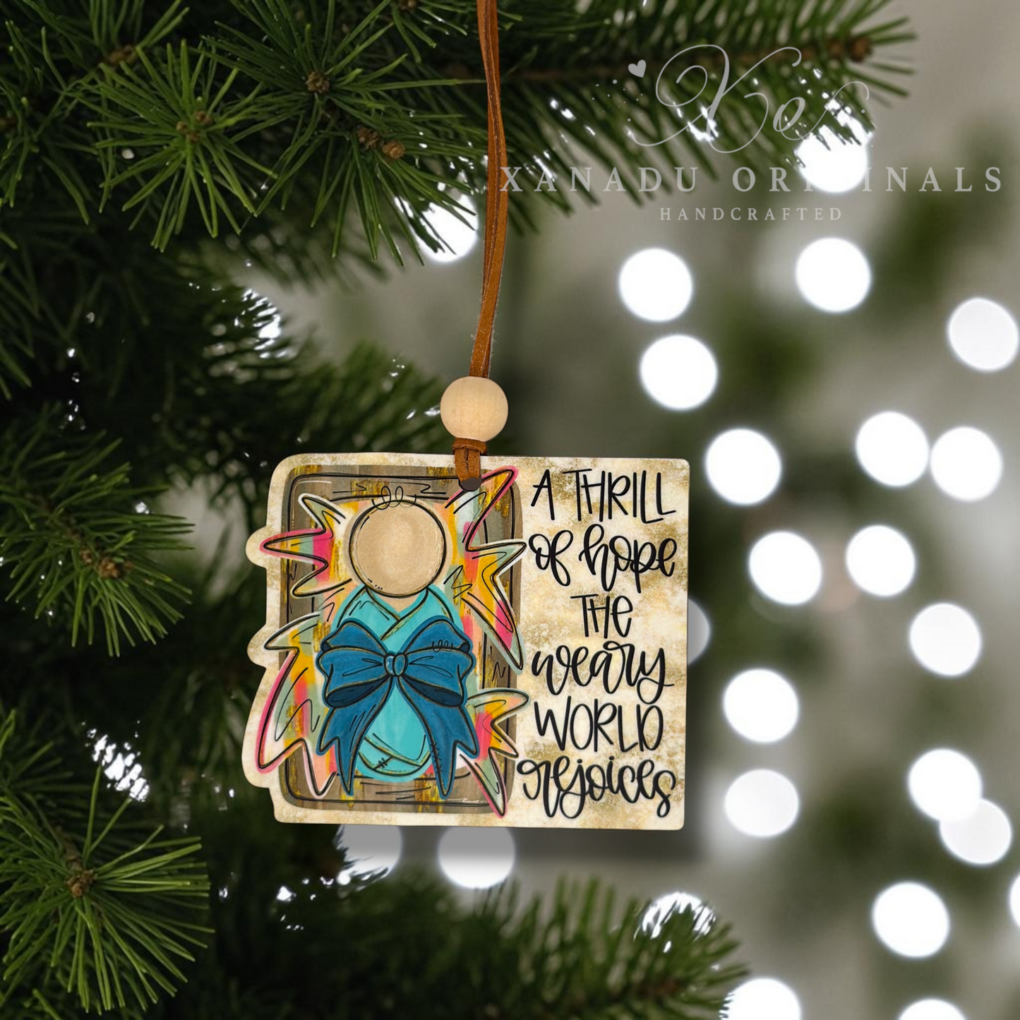 A handcrafted Christmas ornament shaped like an open book, hanging from a pine tree branch. The ornament features a turquoise bow, a gold star, and the text 'A THRILL OF HOPE THE WEARY WORLD rejoices' on a beige background.
