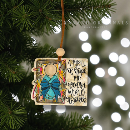 A handcrafted Christmas ornament shaped like an open book, hanging from a pine tree branch. The ornament features a turquoise bow, a gold star, and the text 'A THRILL OF HOPE THE WEARY WORLD rejoices' on a beige background.