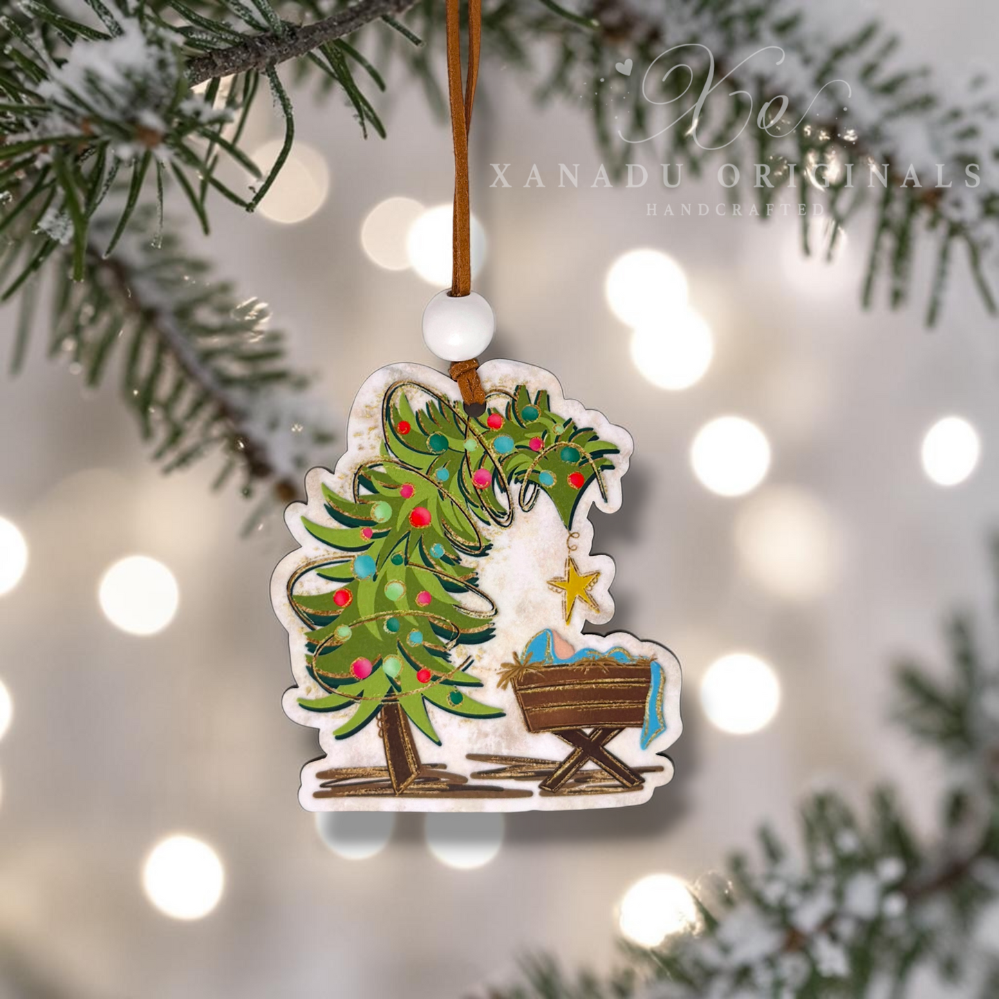 A handcrafted Christmas ornament featuring a green Christmas tree with colorful ornaments and a baby cradle, hanging from a brown string with a white bead. The ornament is shown hanging against a blurred background of pine branches and warm bokeh lights, with the brand name 'Xanadu Originals Handcrafted' visible in the upper right.