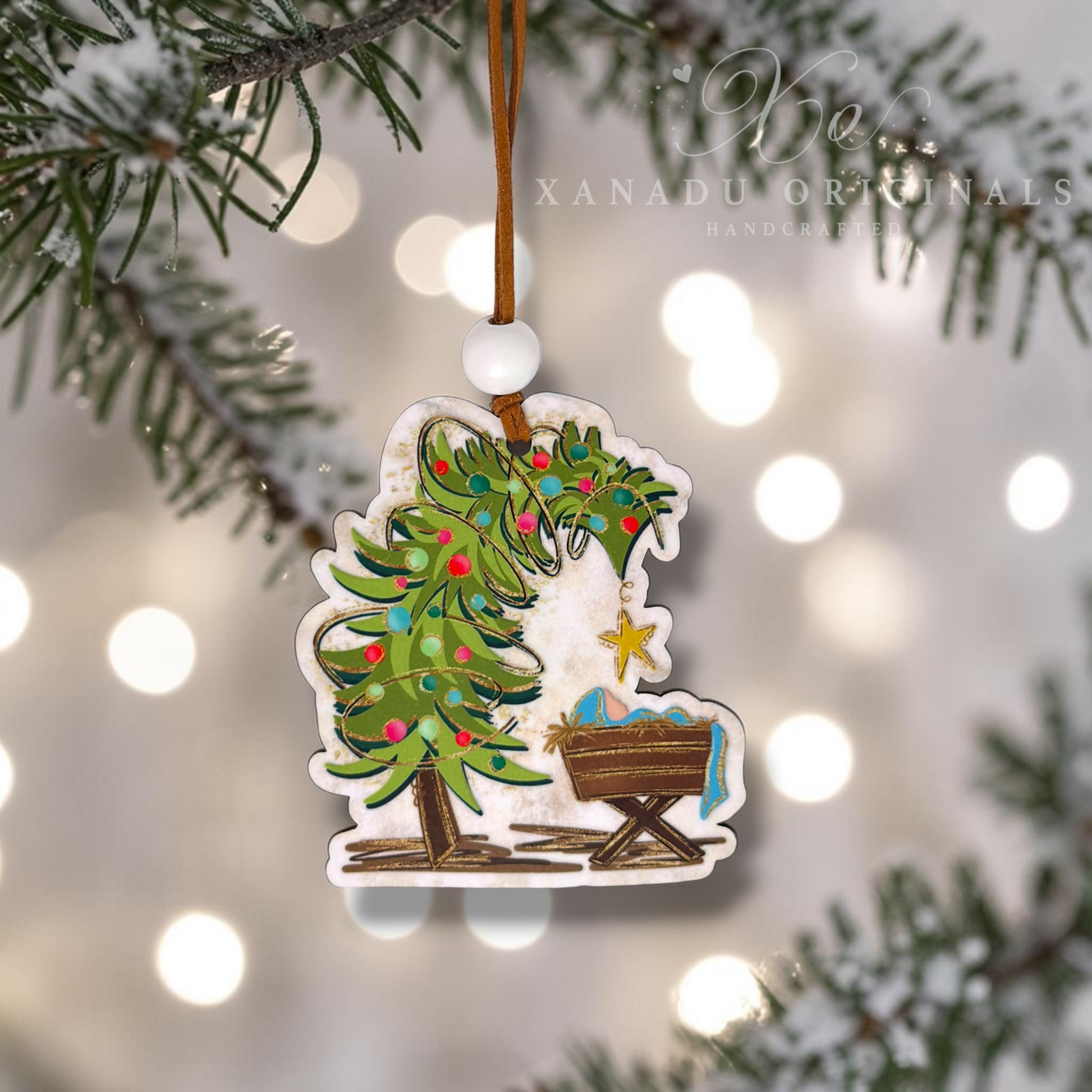 A handcrafted Christmas ornament featuring a green Christmas tree with colorful ornaments and a baby cradle, hanging from a brown string with a white bead. The ornament is shown hanging against a blurred background of pine branches and warm bokeh lights, with the brand name 'Xanadu Originals Handcrafted' visible in the upper right.