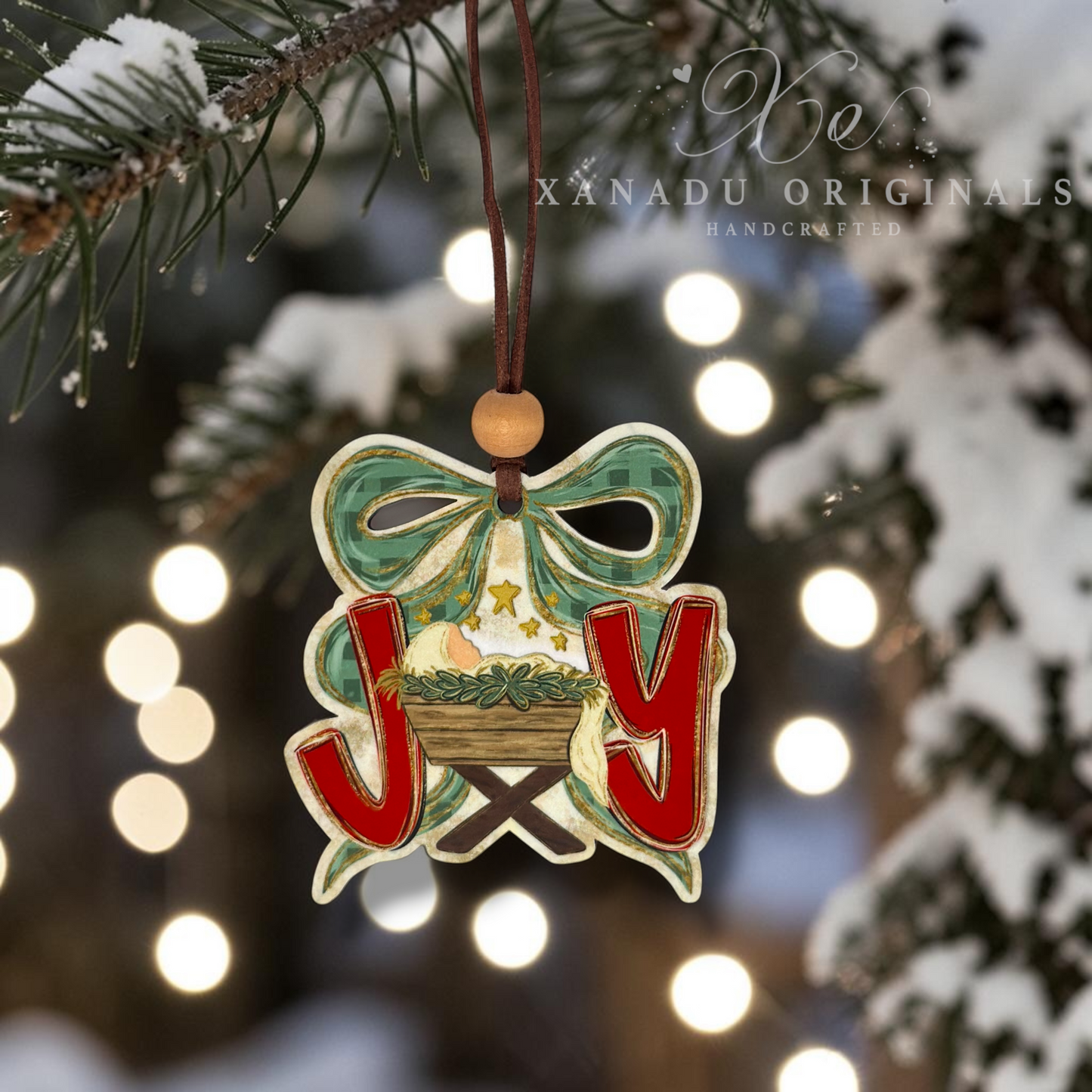A handcrafted Christmas ornament shaped like a nativity scene with the letters J, and O, hanging from a snowy pine branch. The ornament features a green bow, red letters, and a manger scene with a baby Jesus, set against a blurred background of festive lights and snow.