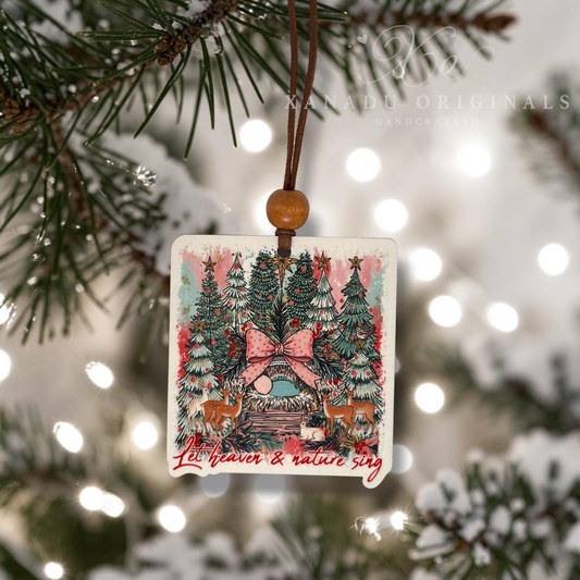 A square Christmas ornament with rounded corners hangs from a brown cord with a wooden bead. The ornament features a colorful winter forest illustration with pine trees, a decorated house, deer, and the phrase 'Let heaven & nature sing' in red script.