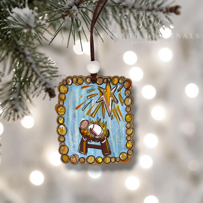 A rectangular hanging Christmas ornament with a painted nativity scene, suspended from a brown cord with a white bead, hanging from a pine branch against a blurred background of warm white bokeh lights.