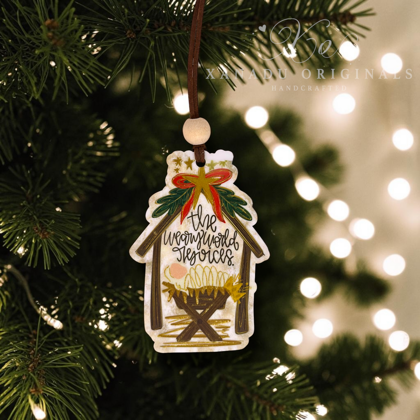 A handcrafted Christmas ornament shaped like a stable, hanging from a brown string on a Christmas tree. The ornament features an illustrated nativity scene with a manger, gold stars, green holly leaves, and the text 'the weary world rejoices'.