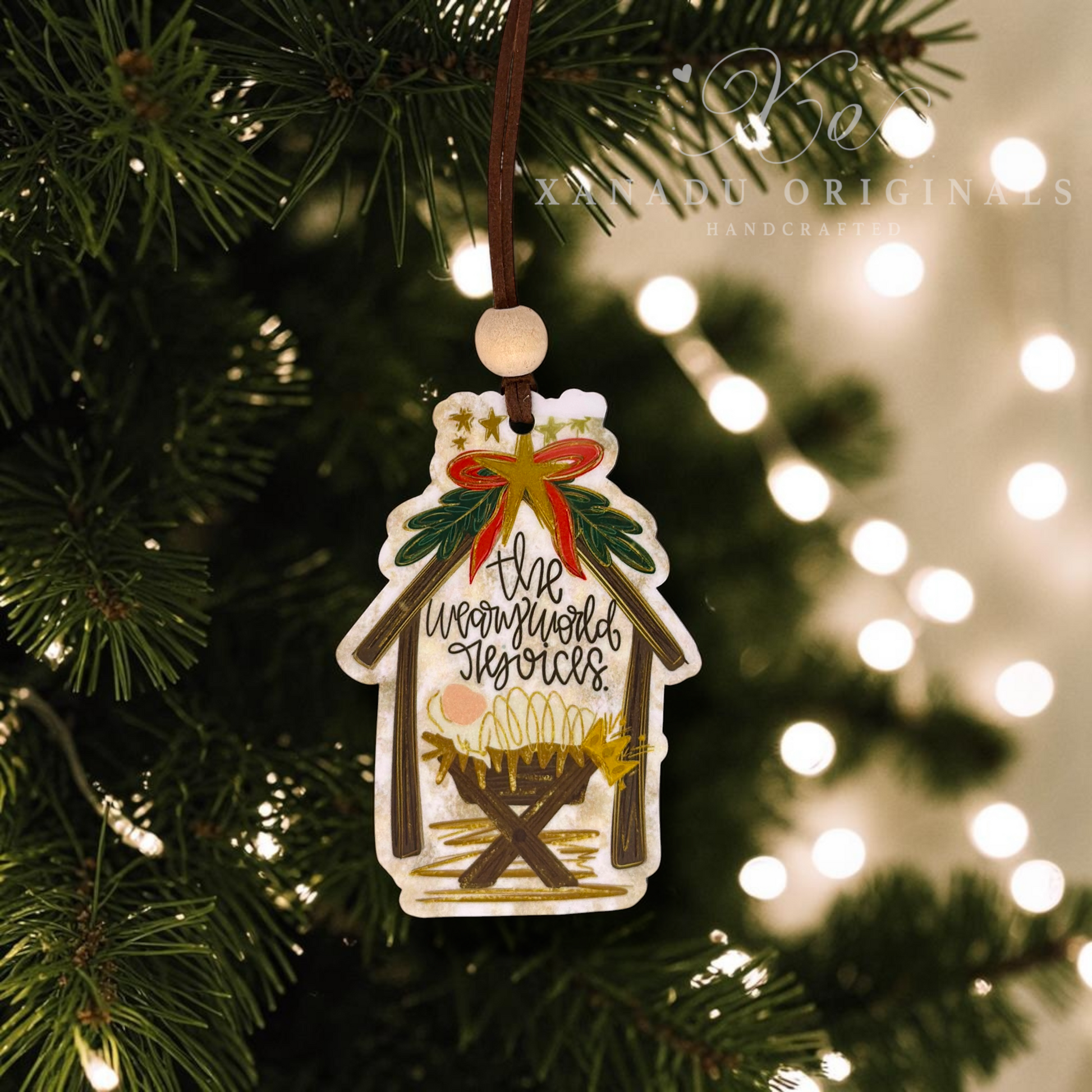A handcrafted Christmas ornament shaped like a stable, hanging from a brown string on a Christmas tree. The ornament features an illustrated nativity scene with a manger, gold stars, green holly leaves, and the text 'the weary world rejoices'.