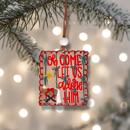 A square wooden Christmas ornament hanging from a brown string and white bead, decorated with red lettering that reads 'Oh Come Let Us adore Him' and a small nativity scene, set against a blurred background of pine branches and warm bokeh lights. Xanadu Originals branding is visible in the top right corner.