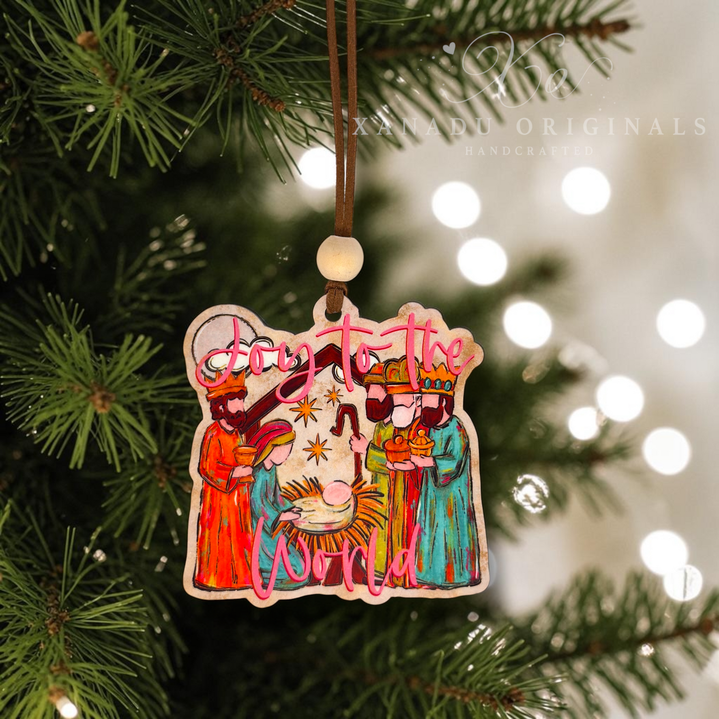 A handcrafted wooden Christmas ornament hanging from a pine tree branch. The ornament features a nativity scene with three wise men, a baby in a cradle, and the words 'Joy to the World' in pink cursive lettering.