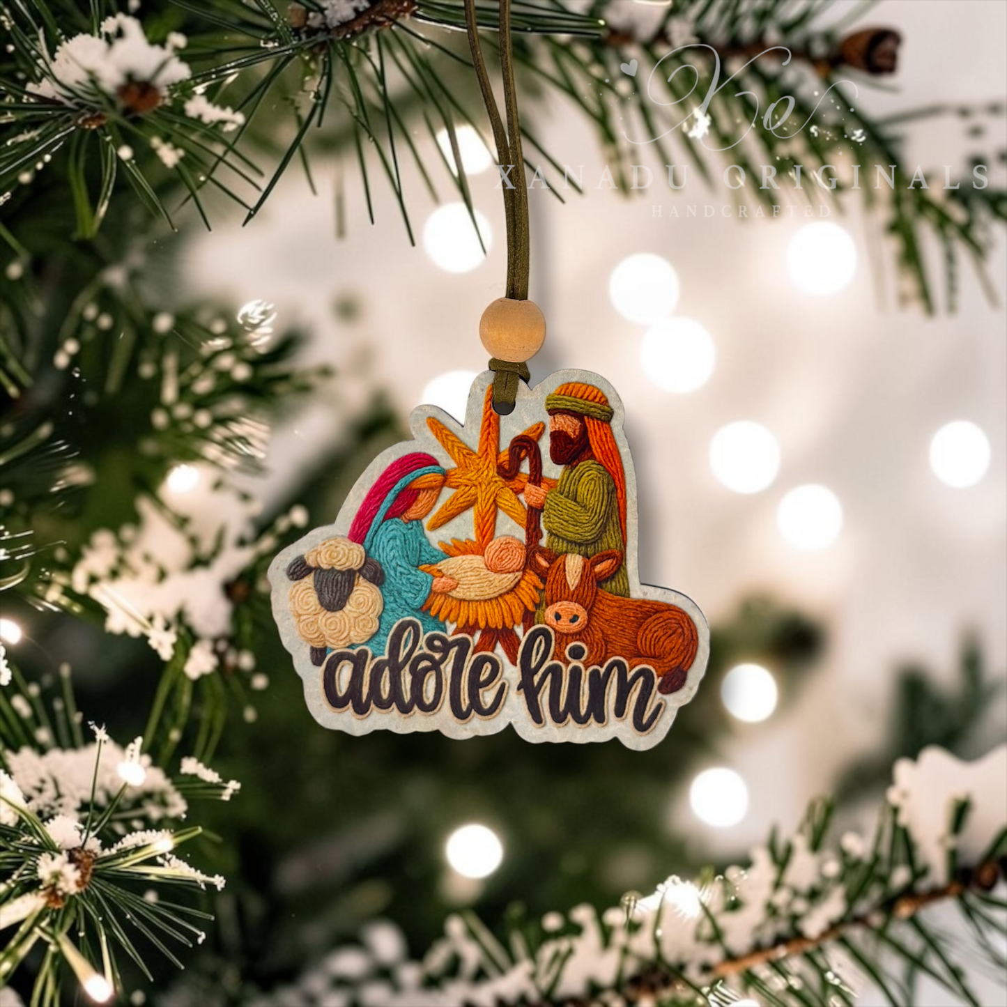 A Christmas ornament shaped like a nativity scene hangs from a tree branch. The ornament features embroidered-style illustrations of Mary, Joseph, baby Jesus, a sheep, and a donkey above the words “adore him,” with a wooden bead and cord for hanging.
