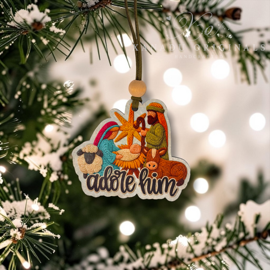 A Christmas ornament shaped like a nativity scene hangs from a tree branch. The ornament features embroidered-style illustrations of Mary, Joseph, baby Jesus, a sheep, and a donkey above the words “adore him,” with a wooden bead and cord for hanging.