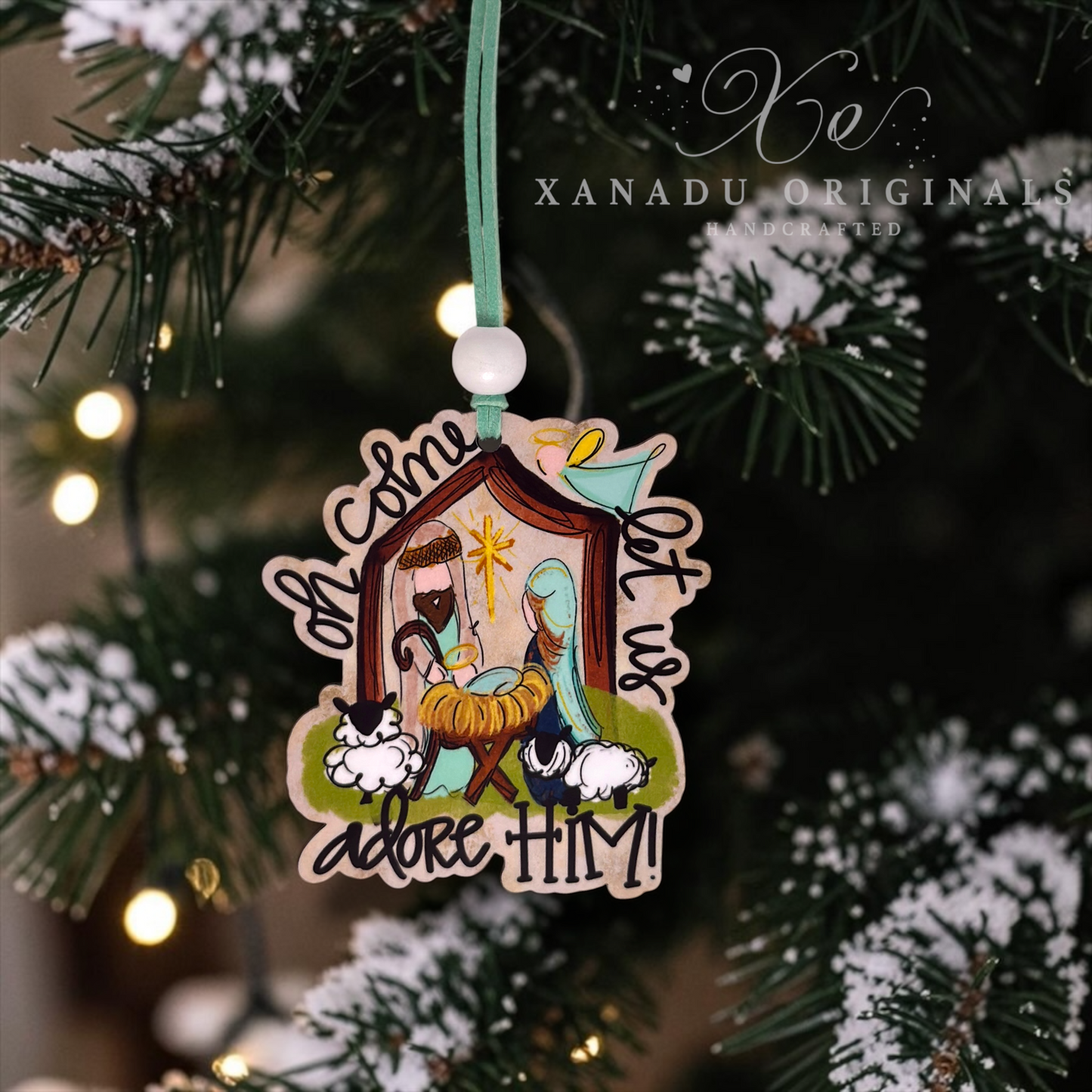 A wooden Christmas ornament featuring a hand-painted nativity scene with Mary, Joseph, baby Jesus in a manger, and sheep, hanging from a green ribbon on a decorated Christmas tree. The text on the ornament reads 'Oh Come Let Us adore HIM!'.