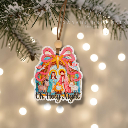 A handcrafted wood Christmas ornament featuring a colorful nativity scene with the text 'Oh Holy Night' at the bottom, hanging from a wooden string under a pine branch with snow, against a warm bokeh background of string lights. A watermark for 'Xanadu Originals Handcrafted' is visible in the upper right.