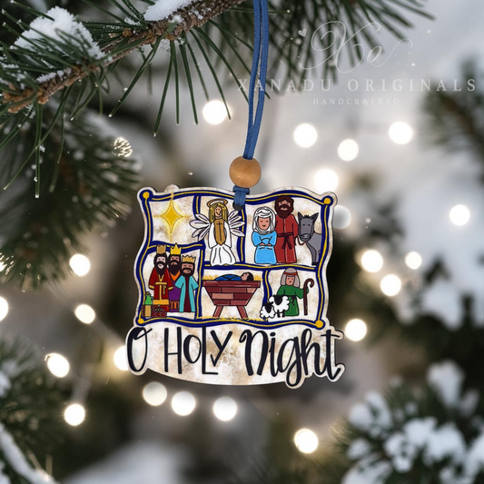 A rectangular wooden Christmas ornament featuring a colorful nativity scene illustration and the words 'O Holy Night' in black script, hanging from a blue string with a wooden bead. The ornament is photographed hanging from a snowy pine branch with blurred holiday lights in the background.