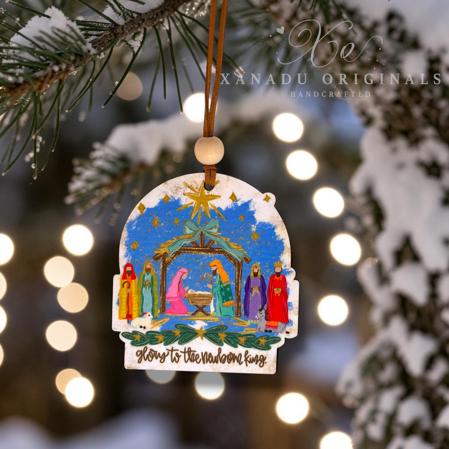 A handcrafted Christmas ornament featuring a colorful nativity scene with the text 'Glory To The Newborn King' at the bottom, hanging from a pine branch with festive bokeh lights in the background.