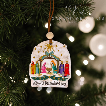 A handcrafted Christmas ornament shaped like a rustic white sign, featuring a painted nativity scene with Mary, Joseph, baby Jesus, and wise men, hanging from a brown string on a Christmas tree branch. The ornament has a weathered wood texture and the phrase 'Glory to the Newborn King' at the bottom.