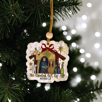 A scalloped wooden Christmas ornament featuring a hand-painted nativity scene with Mary, Joseph, baby Jesus, and animals, hanging from a pine tree branch with a gold ribbon. The ornament is printed with the text 'The Greatest Art Of All MATTHEW 1:21'.