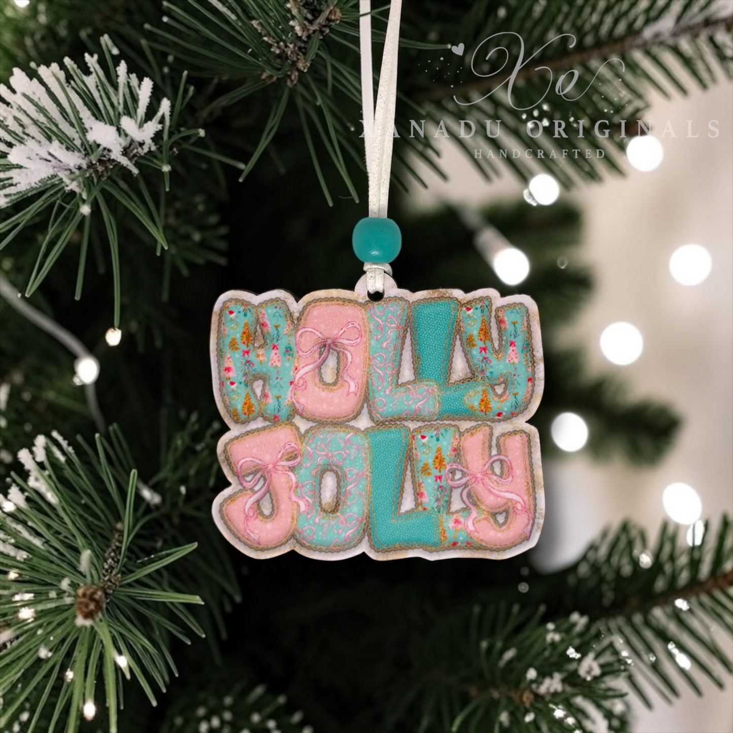A handmade Christmas ornament spelling 'HOLLY JOLLY' in pink and teal icing-like letters, hanging from a white string on a decorated Christmas tree with snowy branches and warm bokeh lights in the background.