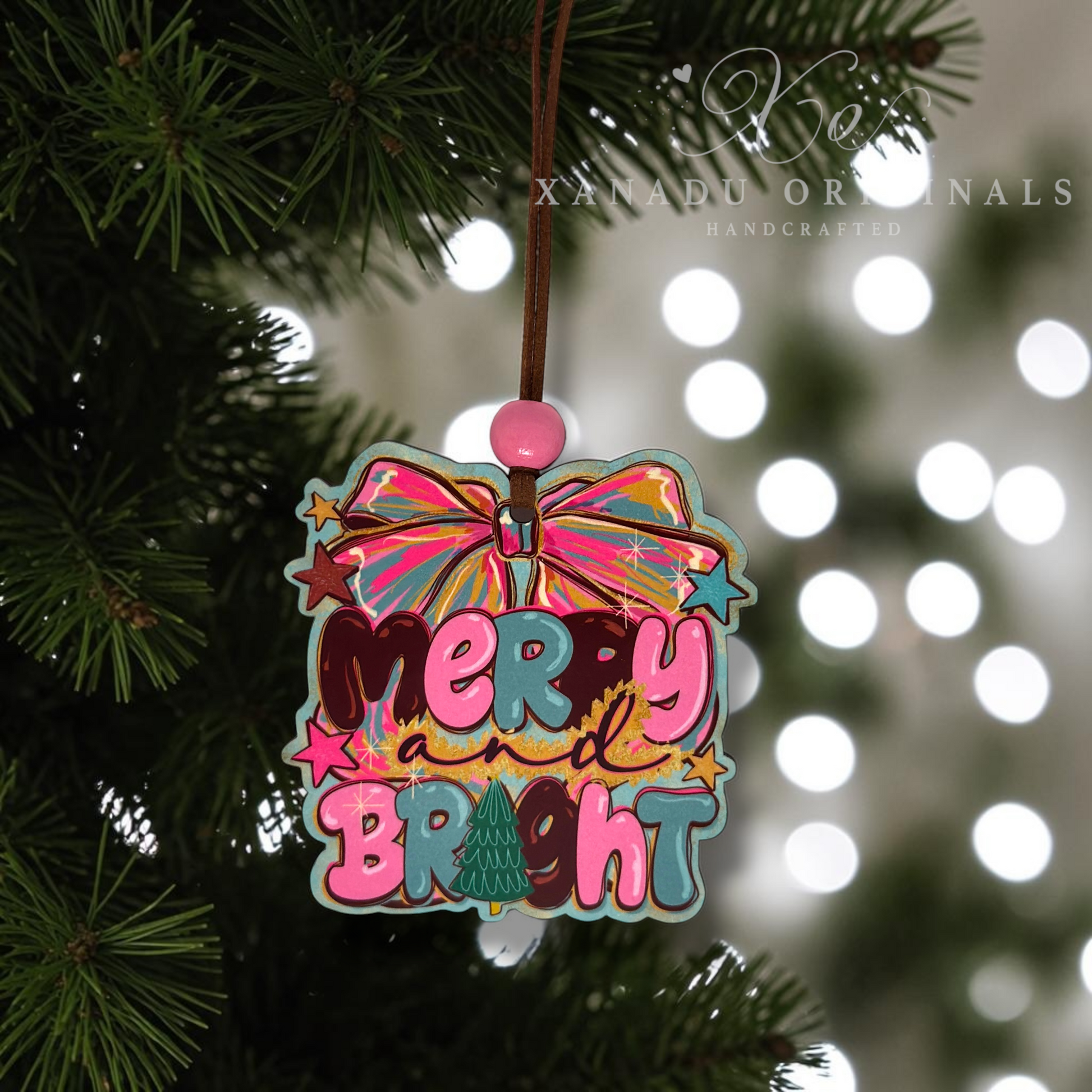 A colorful Christmas ornament shaped like a gift box hanging from a tree branch. The ornament features the words “Merry and Bright” in playful multicolored lettering with a pink bow at the top, surrounded by stars and a teal border.