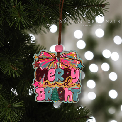 A colorful Christmas ornament shaped like a gift box hanging from a tree branch. The ornament features the words “Merry and Bright” in playful multicolored lettering with a pink bow at the top, surrounded by stars and a teal border.