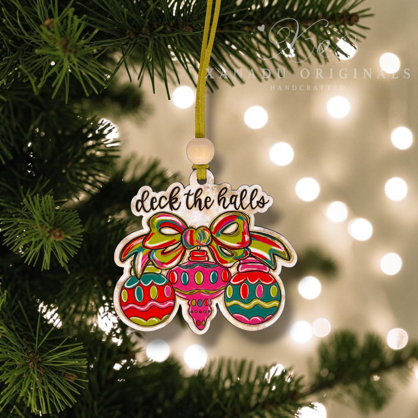 A colorful Christmas ornament hanging from a gold ribbon on a pine tree branch, with blurred holiday lights in the background. The ornament features the phrase 'deck the halls' above three decorated ornaments in bright red, green, yellow, and blue, with a white beaded loop at the top.