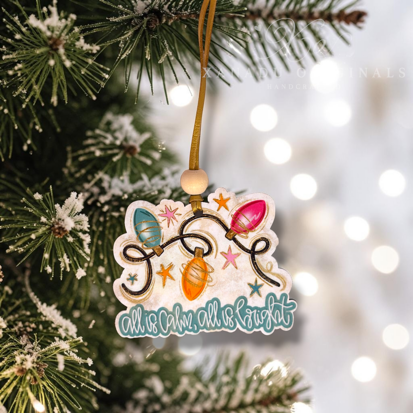 A decorative Christmas ornament hanging from a tree branch. The ornament features a die-cut design with colorful Christmas string lights, stars, and the phrase 'all calm, all night' in teal script.