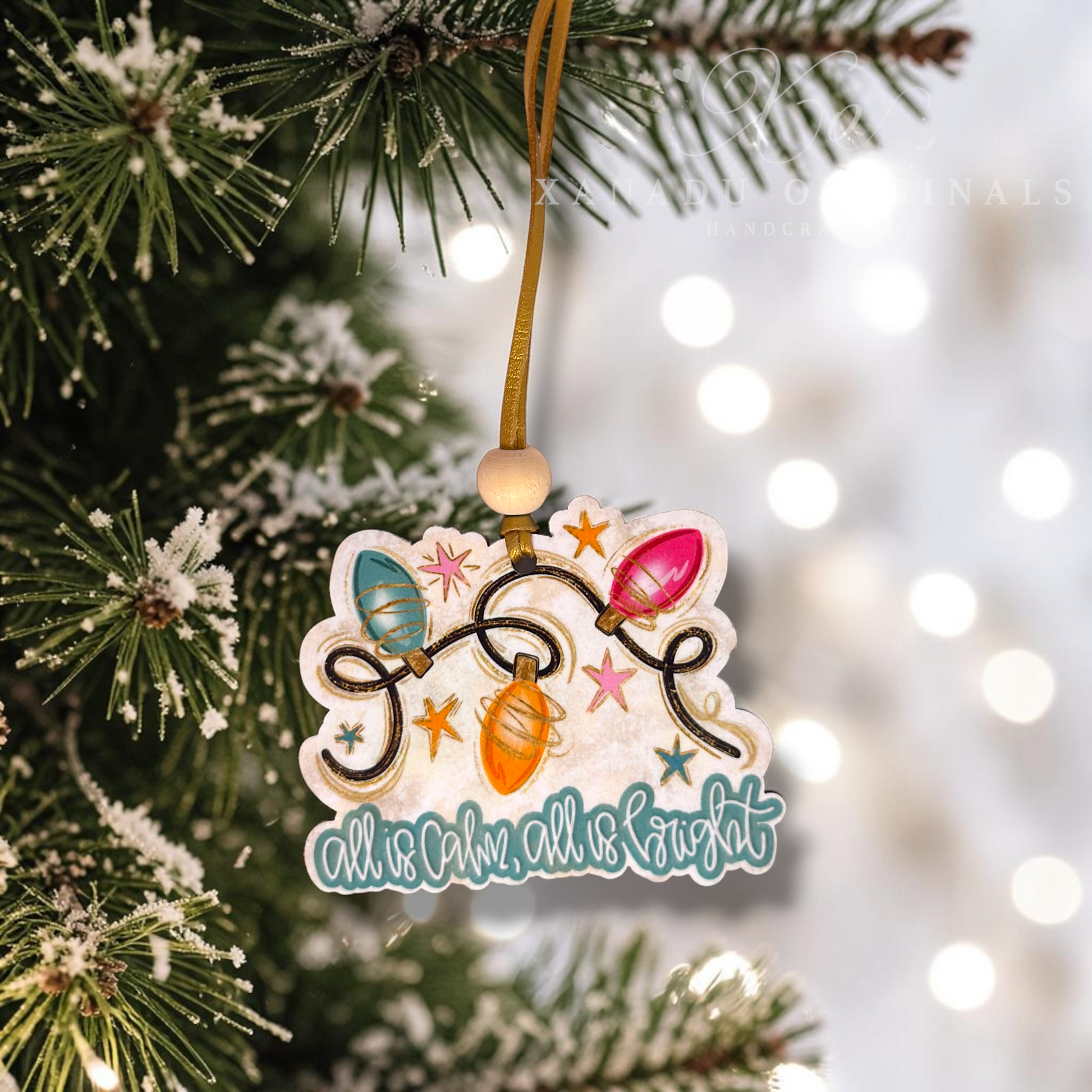 A decorative Christmas ornament hanging from a tree branch. The ornament features a die-cut design with colorful Christmas string lights, stars, and the phrase 'all calm, all night' in teal script.