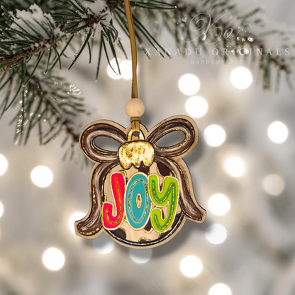 A handcrafted Christmas ornament shaped like a bauble with the word 'Joy' in colorful letters, hanging from a pine branch with warm bokeh lights in the background. The brand name 'Xanadu Originals' is visible as a watermark.
