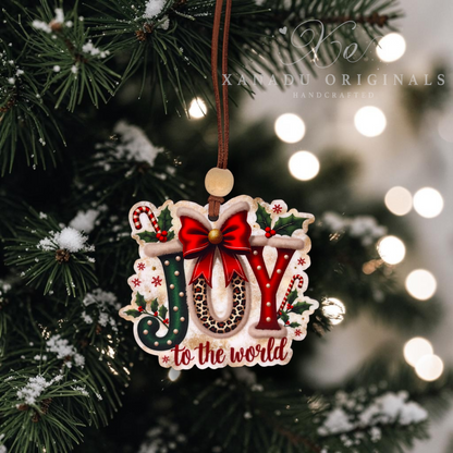 Handcrafted Christmas ornament featuring the word 'JOY' in red and green with a red bow, hanging from a brown cord on a decorated Christmas tree with warm bokeh lights in the background. The ornament has 'to the world' written at the bottom and a watermark for 'Xanadu Originals Handcrafted' is visible.