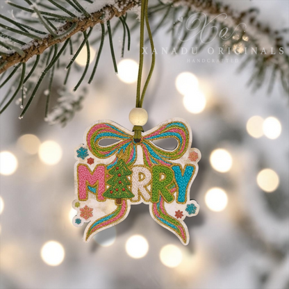 A handcrafted embroidered Christmas ornament shaped like a bow with the word “MERRY” in colorful thread, hanging from a snowy pine branch with warm blurred lights in the background. A faint “XANADU ORIGINALS HANDCRAFTED” watermark appears across the top.