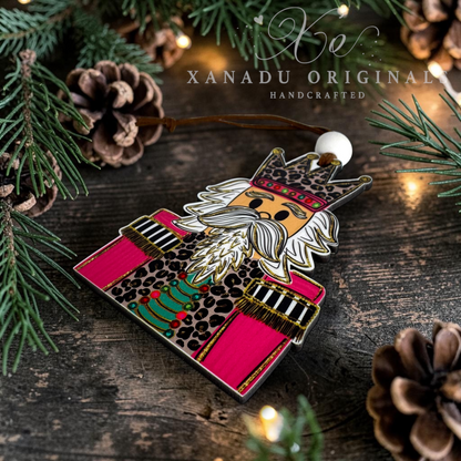 A handcrafted wooden ornament shaped like the Nutcracker, featuring a leopard-print coat, bright pink trousers and a white beard, displayed on a rustic dark wood surface with pine branches, pinecones, and warm string lights. The brand name 'XANADU ORIGINALS HANDCRAFTED' is watermarked at the top.