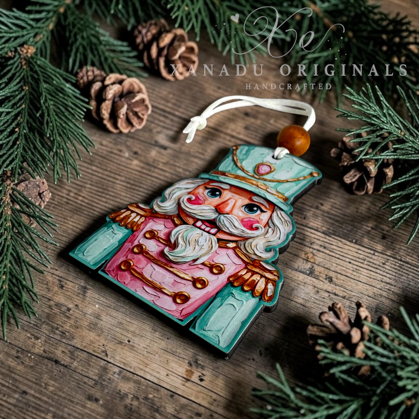 A handcrafted wooden nutcracker ornament by Xanadu Originals, featuring a painted pink and teal design with gold accents, resting on a rustic wooden surface surrounded by pine needles and pinecones.