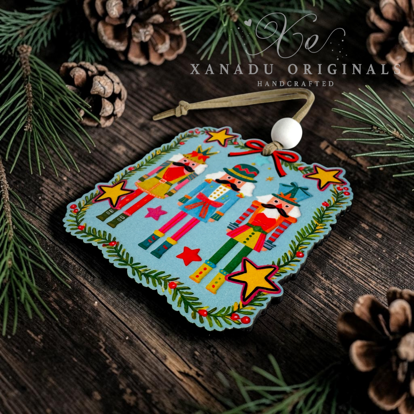 A handcrafted wooden ornament featuring three nutcracker figures on a light blue background, surrounded by yellow stars and green garland details. The ornament is decorated with a white bead and a wooden loop, set against a rustic dark wood background with pine needles and pinecones.