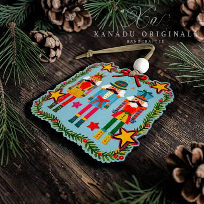 A handcrafted wooden ornament featuring three nutcracker figures on a light blue background, surrounded by yellow stars and green garland details. The ornament is decorated with a white bead and a wooden loop, set against a rustic dark wood background with pine needles and pinecones.