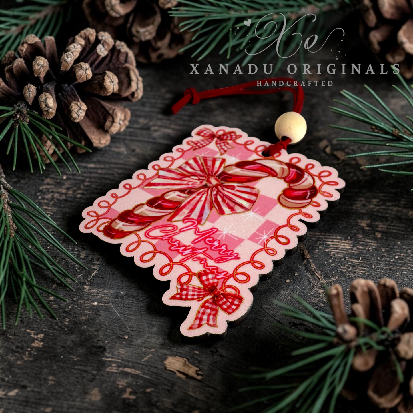 A handcrafted Christmas ornament shaped like a gift tag, featuring a red and pink plaid pattern with candy cane illustrations, red script text, and a red hanging cord with a small wooden bead. The ornament is styled on a dark rustic wooden surface with pine needles and pinecones, and the brand name 'XANADU ORIGINALS' is visible at the top.