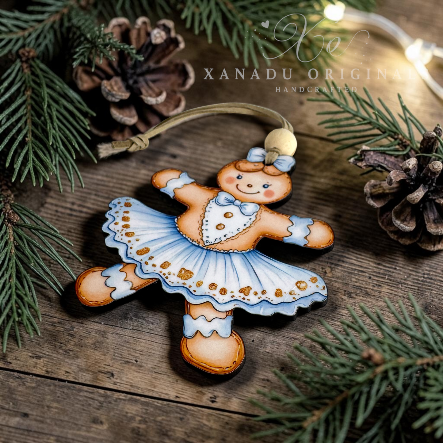 A handcrafted wooden gingerbread ornament shaped like a dancing gingerbread woman in a blue tutu, displayed on a rustic wooden surface with pine branches and pinecones. A watermark for 'XANADU ORIGINAL HANDCRAFTED' is visible in the upper right.