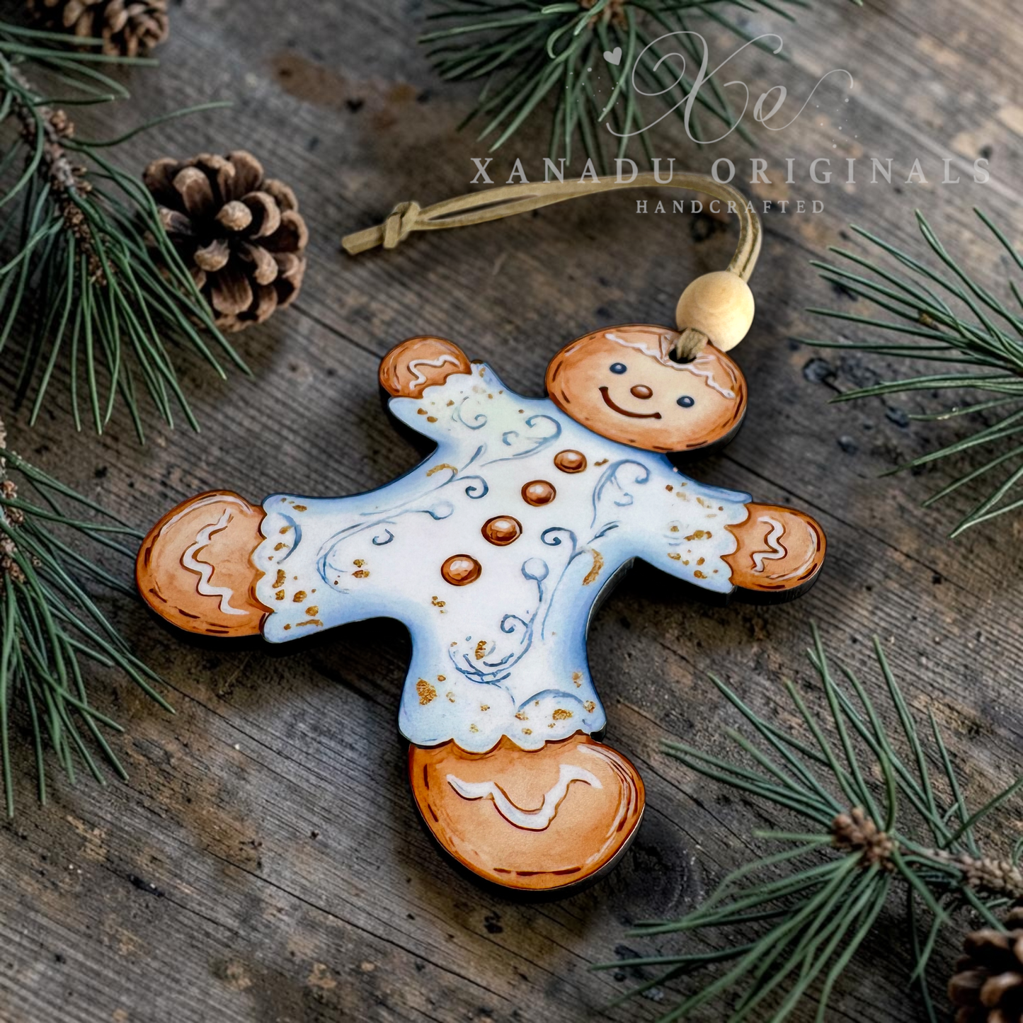 Handcrafted gingerbread man Christmas ornament with a white icing body, brown painted joints and head, hanging on a rustic wooden surface with pine branches and pinecones. XANADU ORIGINALS branding is visible.