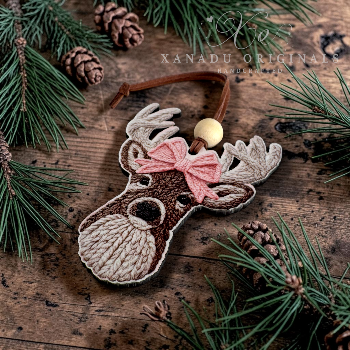 A custom-cut wood ornament of a faux knit textured deer, featuring a pink bow, brown and beige fur details, and a wooden bead on a faux leather loop, displayed on a rustic wooden surface with pine branches and pinecones. A watermark for 'XANADU ORIGINALS HANDCRAFTED' is visible at the top.