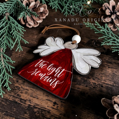 Handcrafted Christmas angel ornament with a red body, white wings, and the words 'the light remains' in white script, displayed on a rustic wooden surface with pine branches and pinecones. Xanadu Originals branding is visible.