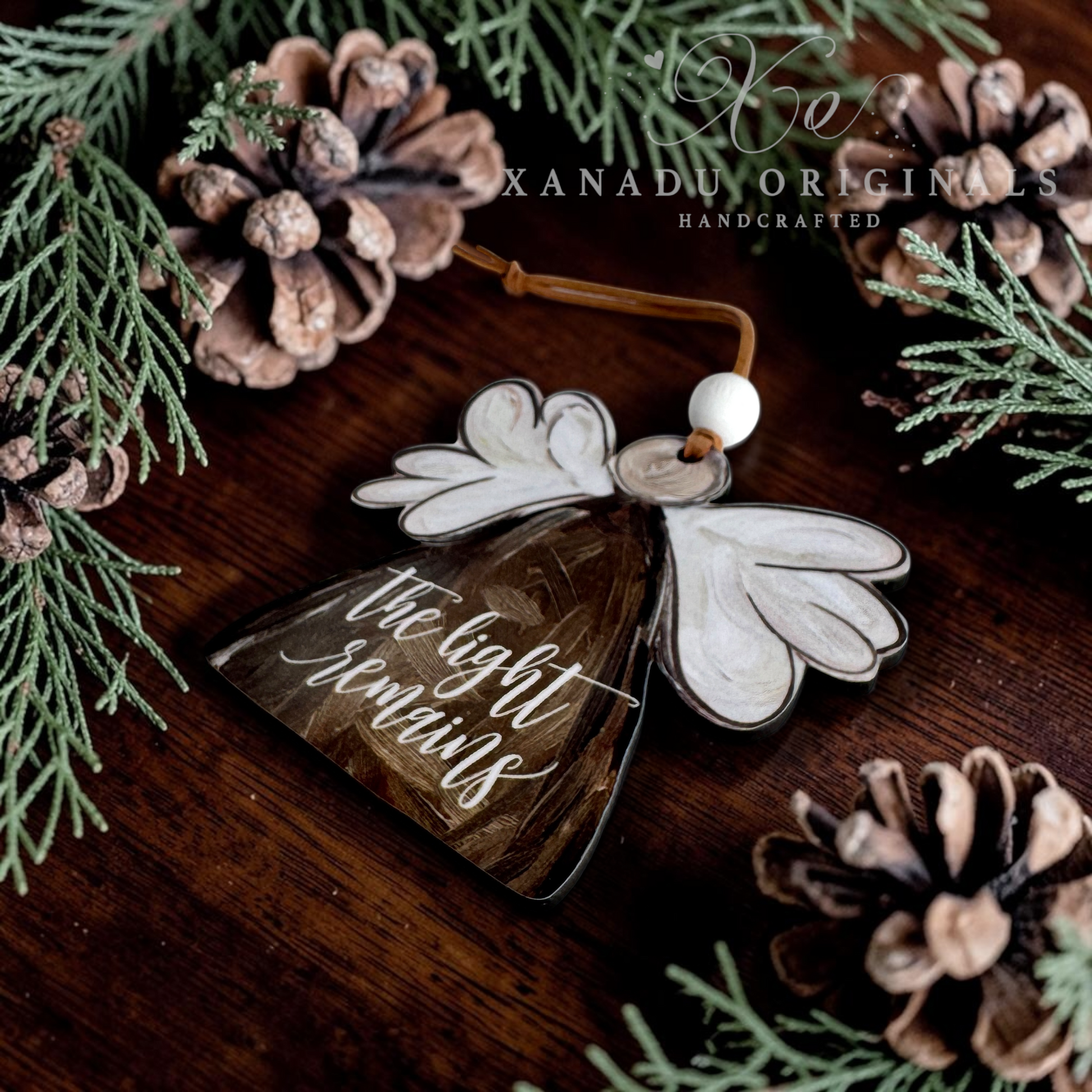A handcrafted wood angel ornament with white wings and a brown body engraved with 'the light remains', hanging on a dark wooden surface surrounded by pinecones and green pine branches. The brand name 'XANADU ORIGINALS' is visible as a watermark at the top.