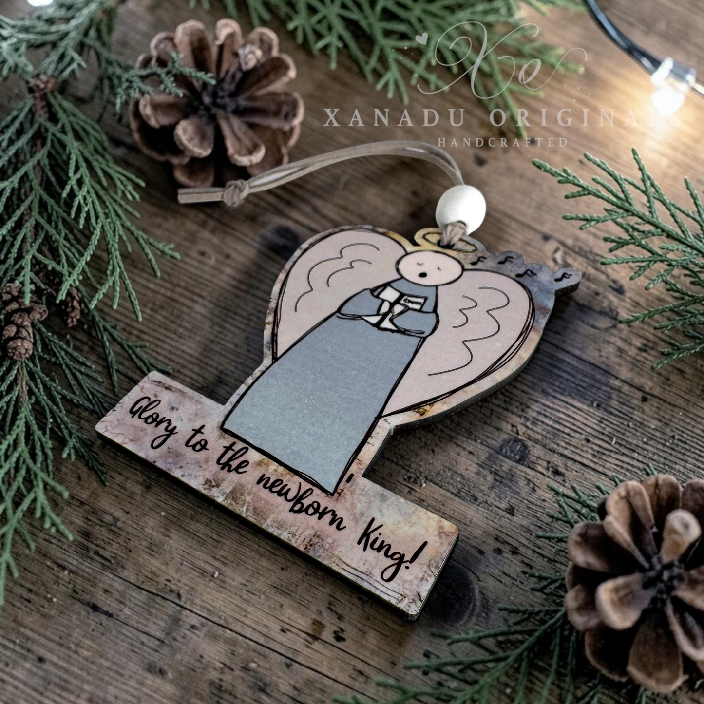 A handcrafted wooden Christmas ornament shaped like an angel with the text 'Glory to the newborn King!' on a rustic wooden surface, surrounded by pine branches, pinecones, and warm string lights. A watermark for 'XANADU ORIGINAL HANDCRAFTED' is visible.