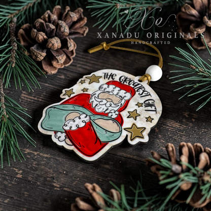 A handcrafted wooden Christmas ornament featuring Santa Claus in a red suit and baby Jesus, surrounded by gold stars and the text 'THE GREATEST GIFT'. The ornament is displayed on a dark wooden surface with pine needles and pinecones, with a watermark for 'XANADU ORIGINALS' at the top.