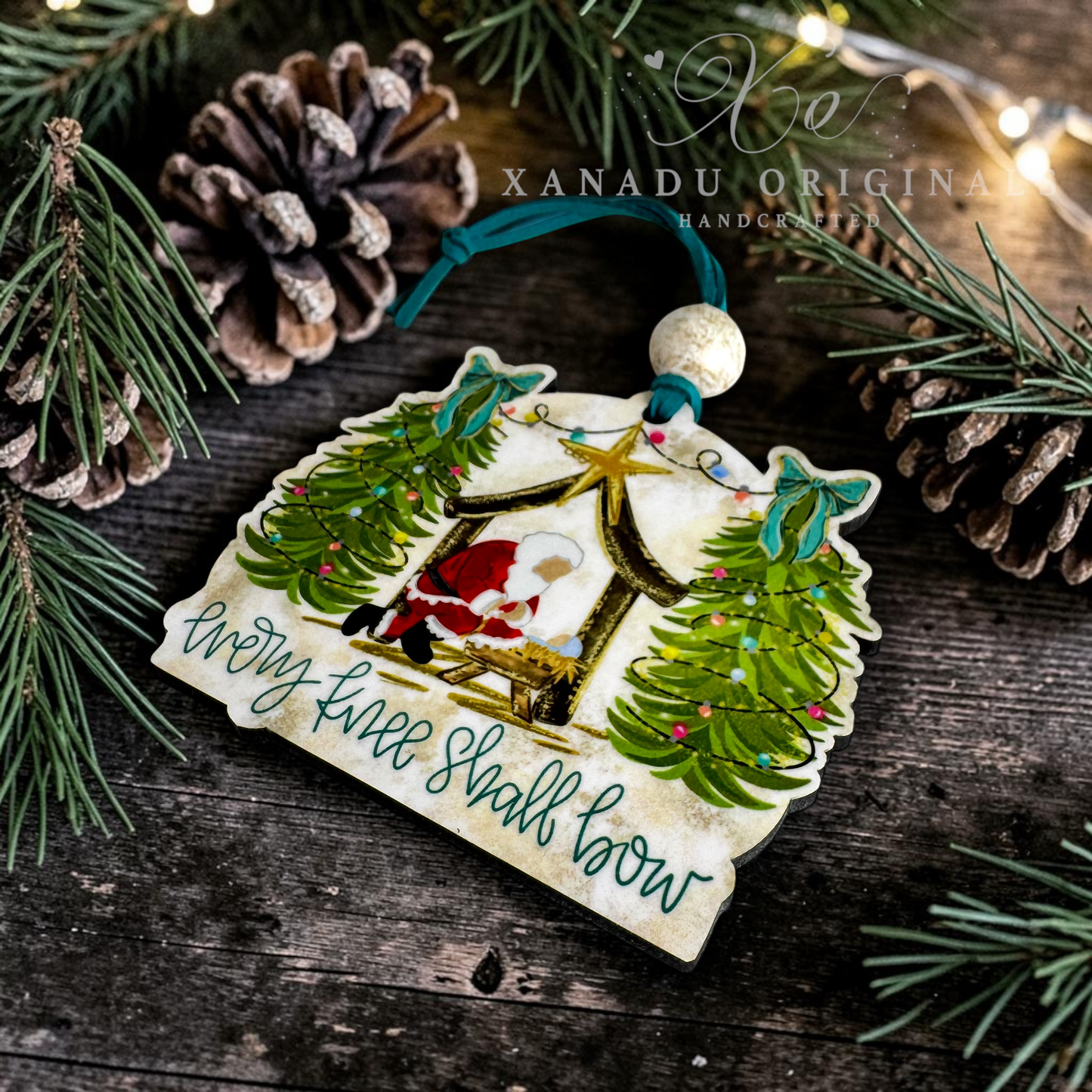 Handcrafted Christmas ornament featuring Santa in a sleigh between decorated trees, with the text 'Every Tree Shall Bow' and a wooden bead hanger, styled on a rustic wooden surface with pine branches and pinecones.
