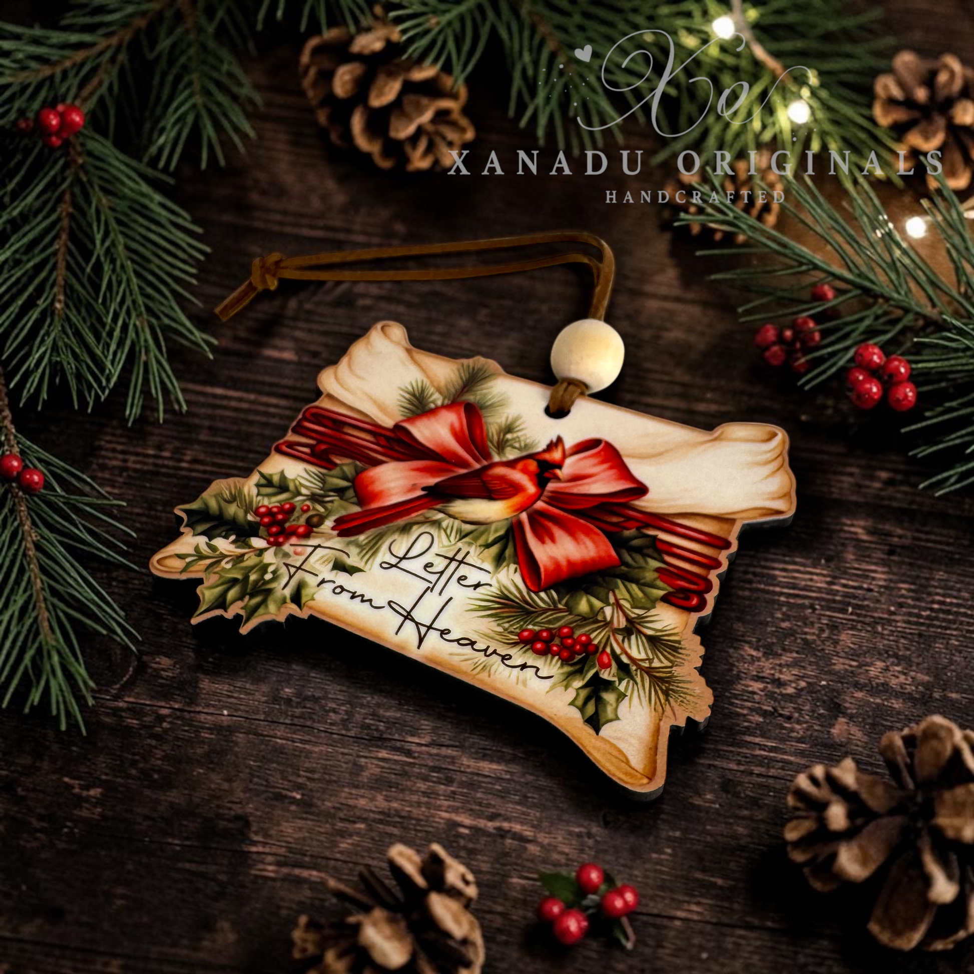 A handcrafted Christmas ornament shaped like a letter addressed 'From Heaven,' decorated with a red bow, green pine branches, holly, and red berries, hanging on a dark wooden surface with pinecones and evergreen branches around it. The text 'Xanadu Originals Handcrafted' appears at the top.