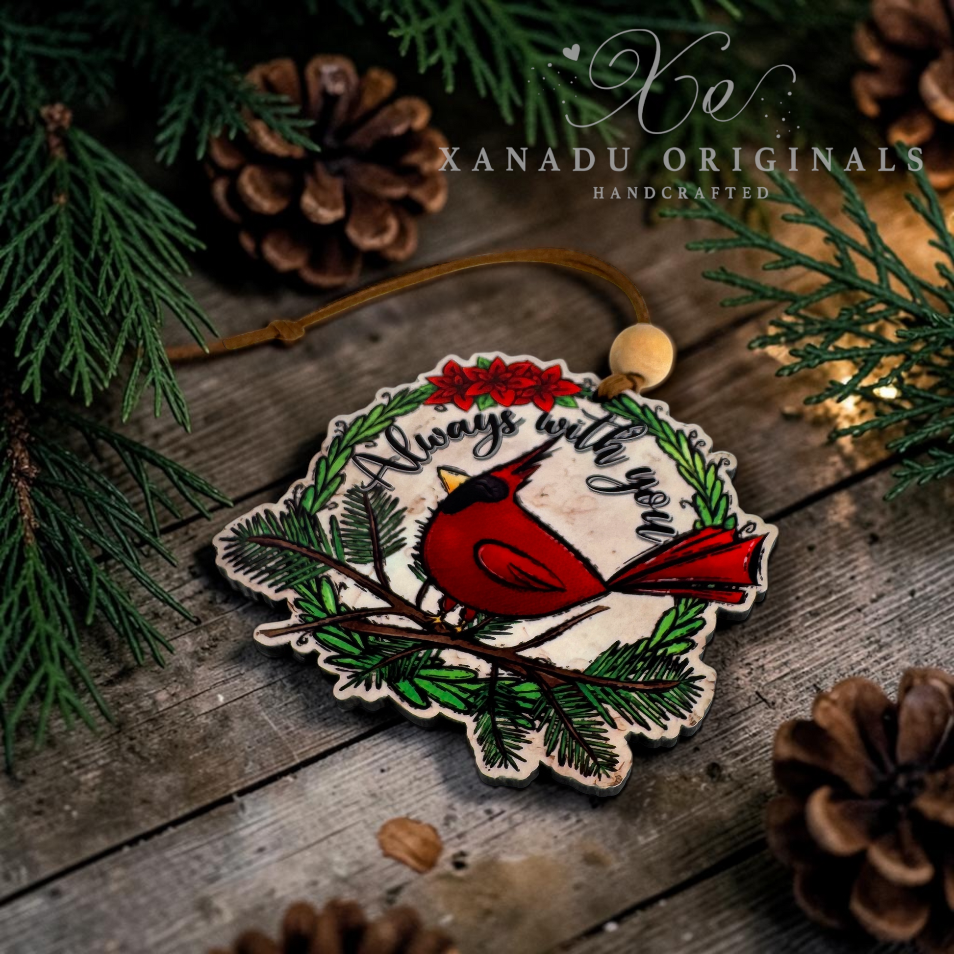 A handcrafted wooden Christmas ornament featuring a red cardinal perched on a branch with green pine needles, red berries, and the phrase 'Always with you' in white script. The ornament is displayed on a rustic wooden surface surrounded by pine branches, pinecones, and warm string lights, with the brand name 'XANADU ORIGINALS' visible at the top.