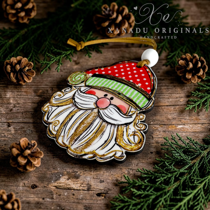 A handcrafted Christmas ornament shaped like Santa's face, featuring a red hat with white polka dots, a green striped band, a white beard with gold glitter accents, and a gold hanging loop. The ornament is displayed on a rustic wooden surface surrounded by pine needles and pinecones, with the brand name 'XANADU ORIGINALS' visible at the top.