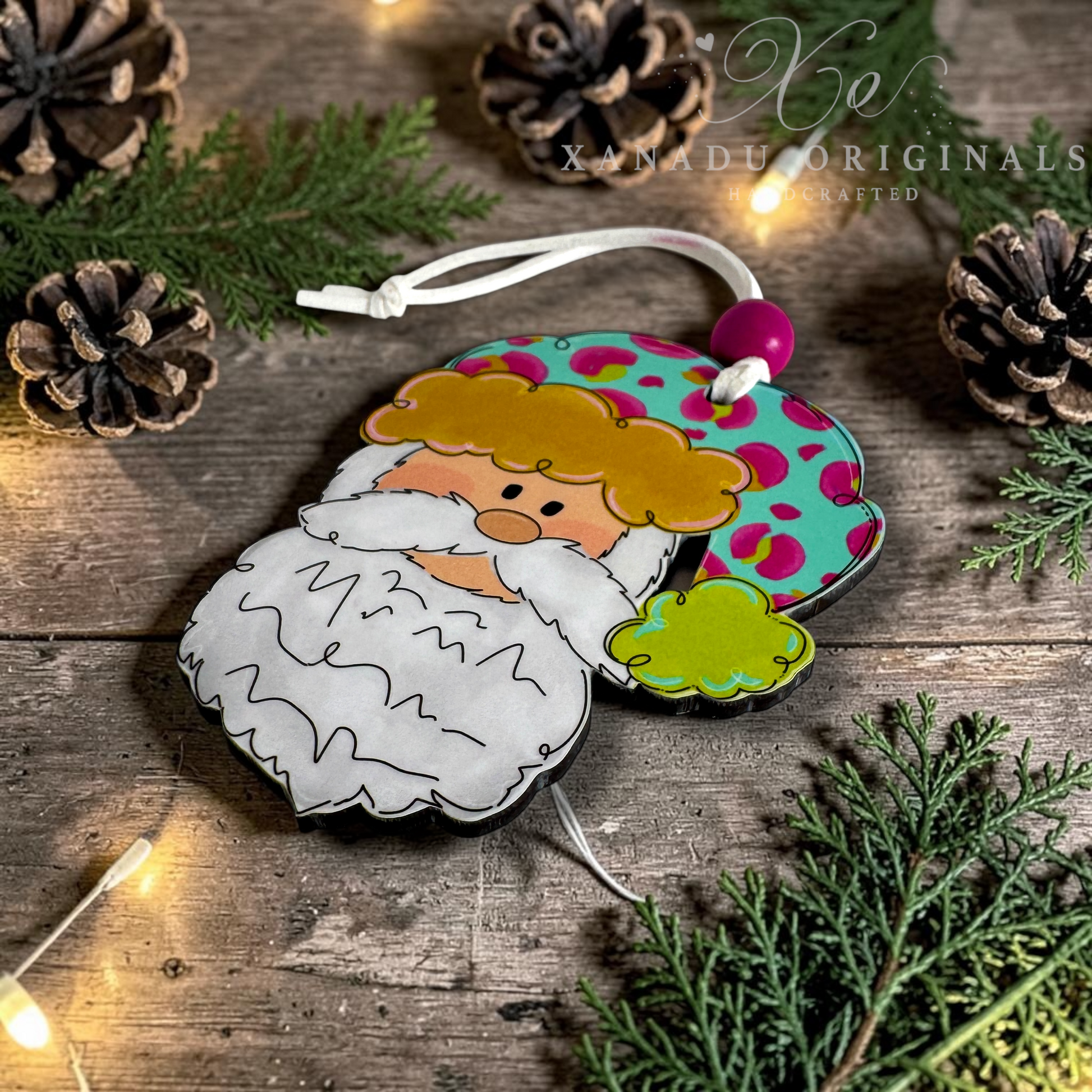 A hand holds a colorful Christmas ornament shaped like a stylized Santa face with a white beard, yellow hair, and a teal hat with pink spots. The background shows holiday greenery, ornaments with a watermark reading XANADU ORIGINALS.