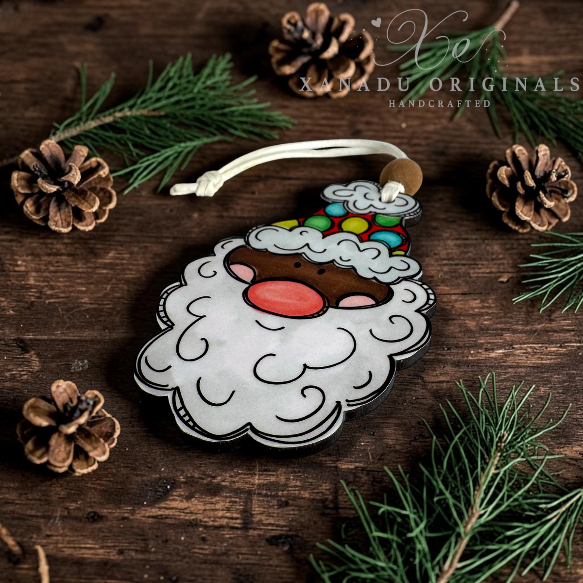 A dark skin tone Santa Claus wood ornament with a colorful patterned hat lying flat on a rustic dark wooden tabletop surrounded by pine cones and evergreen branches.