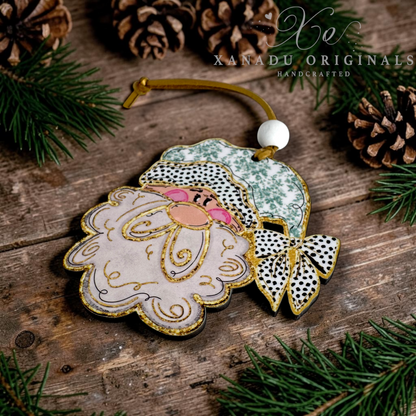 A handcrafted Christmas ornament shaped like Santa's face, featuring a pale pink beard with gold glitter outlines, a green and white patterned hat, black-and-white spotted bow details, and a gold hanging loop. The ornament is styled on a rustic wooden surface with pine branches and pinecones, with the brand name 'XANADU ORIGINALS' visible in the upper right.